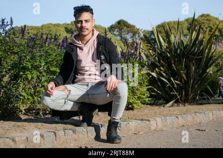 young fashionable homosexual latino man with mustache and short hair, wears pink sweater and black jacket, crouching posing looking at camera in park Foto Stock