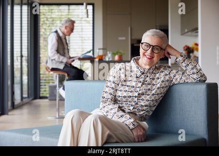 Ritratto di una donna anziana sorridente seduta sul divano a casa con un uomo sullo sfondo Foto Stock