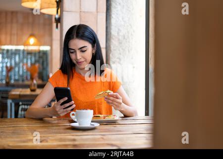 Una giovane donna sorridente che usa uno smartphone e si gusta il cibo in un bar Foto Stock