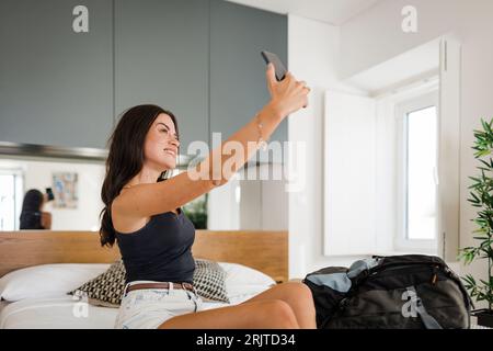 Giovane donna felice che scatta selfie attraverso lo smartphone in camera da letto Foto Stock