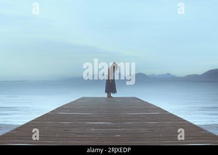 una donna solitaria in piedi su un molo vicino al mare si lascia trasportare dalle emozioni in un'atmosfera blu e surreale Foto Stock