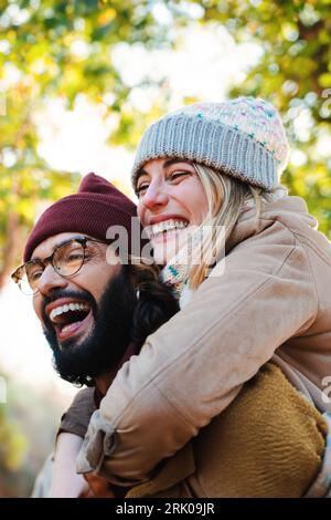 Vertical close up portrait of lovely couple hugging. Young bearded man with glasses giving his blonde cute girlfriend a piggyback ride. Pair of happy friends having fun outside at weekend trip. High quality photo Stock Photo