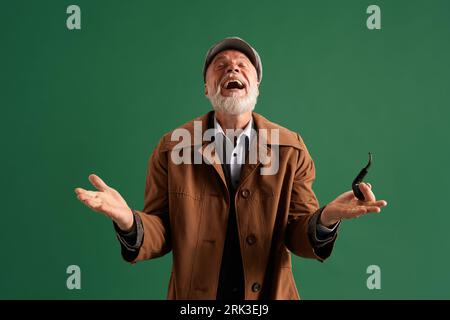 Portrait of positive, contented and happy man, detective wearing vintage hat, elegance suit with pipe in mouth on green color background. Stock Photo