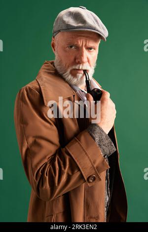 Portrait of confident and strict man, detective wearing vintage hat, elegance suit with pipe in mouth looking at camera on green background. Stock Photo