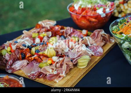 Tavola di legno con salumi a fette, olive e physalis, oltre ad altri spuntini sul tavolo durante il buffet in giardino Foto Stock