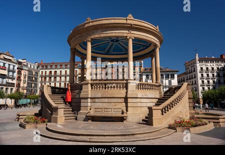 Pamplona, Spagna - 2 agosto 2022: Donna che indossa un abito rosso accanto al monumento Padiglione in Piazza del castello nella città vecchia di Pamplona Spagna. Foto Stock