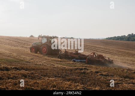 Un trattore moderno con un pesante erpice a disco trainato ara un campo di grano al tramonto. Foto Stock