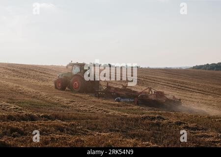 Un trattore moderno con un pesante erpice a disco trainato ara un campo di grano al tramonto. Foto Stock