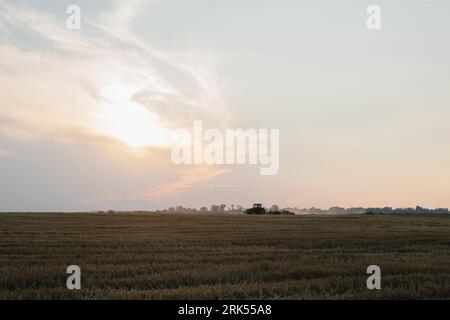 Un trattore moderno con un pesante erpice a disco trainato ara un campo di grano al tramonto. Foto Stock