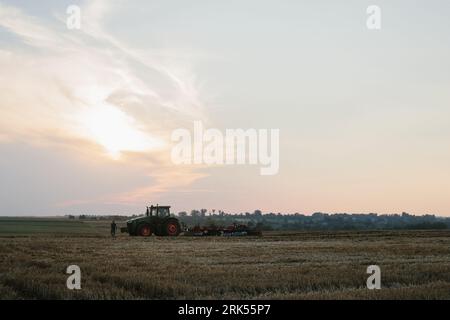 Un trattore moderno con un pesante erpice a disco trainato ara un campo di grano al tramonto. Foto Stock