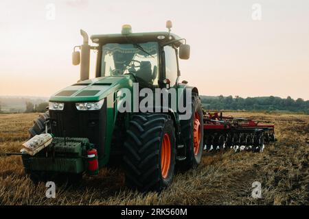 Foto ravvicinata di un moderno trattore verde con un aratro a disco rosso che lavora con il GPS per l'agricoltura di precisione. Foto Stock