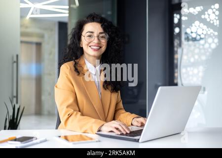 Ritratto di una donna d'affari matura di successo all'interno dell'ufficio sul posto di lavoro, un capo dell'america latina che sorride e guarda la fotocamera utilizzando un notebook al lavoro, soddisfatta dei risultati finanziari ottenuti. Foto Stock