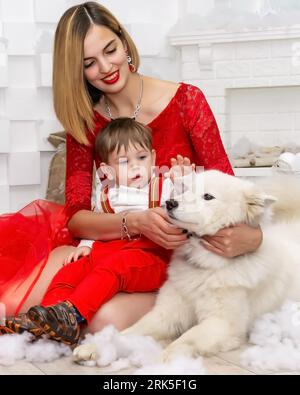 La famiglia accarezza un cane. Ritratto fotografico di famiglia. Mamma e suo figlio e un cane bianco in abiti rossi celebrano i Chistmas, Capodanno. Felice giovane madre, lei Foto Stock