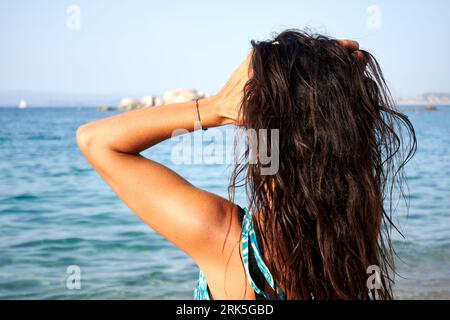A young woman standing on the beach near the shoreline, wearing a bikini top and gazing out to sea Stock Photo