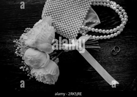 A grayscale shot of wedding flowers, a bag and marriage rings on a wooden background. Stock Photo