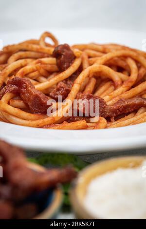 Bucatini fatti in casa all'Amatriciana, guance di manzo cotte su pasta, formaggio e salsa di pomodoro in un recipiente bianco Foto Stock