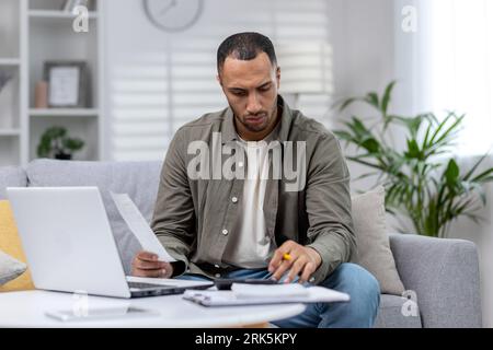 L'uomo latino-americano è concentrato a lavorare da remoto a casa con un laptop e con documenti. Foto Stock