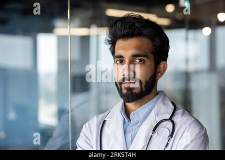 Foto in primo piano. Ritratto di un giovane medico indiano, stagista, assistente che guarda seriamente la macchina fotografica in un cappotto bianco nella clinica. Foto Stock