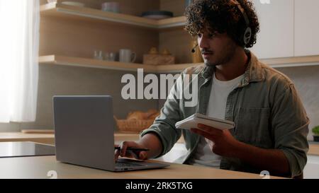 Indian man in headset look app pc browsing typing laptop write ideas questions notes review research sit at home kitchen. Arabian guy learn language Stock Photo