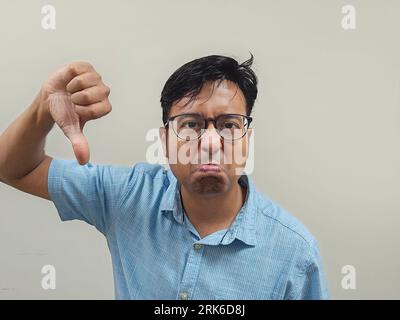 An Indian male wearing glasses and a blue shirt expressing his dissatisfaction by making a thumb down gesture against a gray background Stock Photo