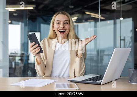 Una giovane donna felice sta lavorando in ufficio a un tavolo con un laptop e tenendo in mano un telefono cellulare. Piacevolmente scioccata e felice, diffonde gli occhi e guarda la telecamera con un sorriso. Foto Stock