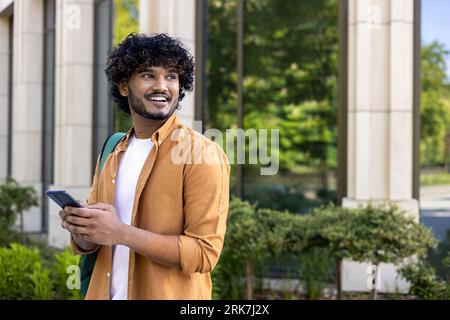 Un giovane indiano con uno zaino e un telefono in mano si trova su una strada cittadina e guarda con un sorriso. viaggi in attesa di incontrarsi. Foto Stock
