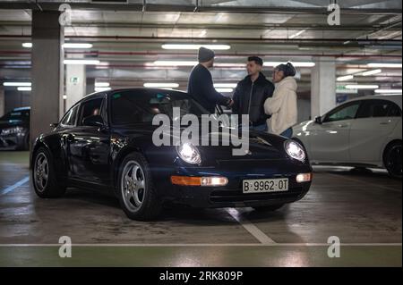 Vista frontale di una classica auto sportiva tedesca, la Porsche 993 di colore nero parcheggiata in garage con alcuni ragazzi Foto Stock