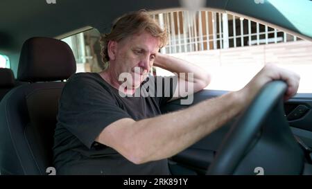 Senior man struggles with life's difficulties inside car parked in street. Desperate older caucasian person suffering in quiet despair Stock Photo