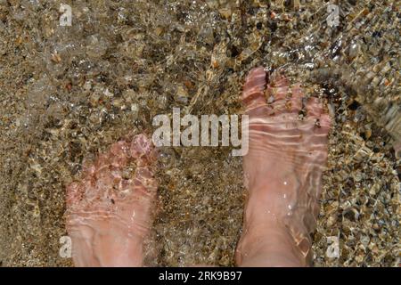 Barefoot women's feet close-up in clear water on the sandy bottom. The glare of the sun on the water. Stock Photo