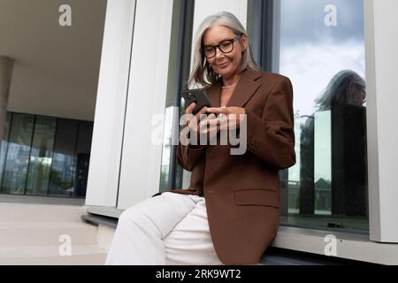 60 anni donna d'affari con i capelli grigi, vestita con stile, controlla i messaggi sulla posta al telefono Foto Stock