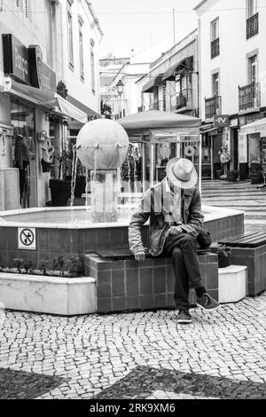 Lagos, Algarve, Portogallo - 20 ottobre 2022: Uomo con cappello bianco seduto su una fontana in una piazza di Lagos Foto Stock