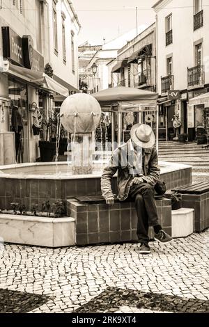 Lagos, Algarve, Portogallo - 20 ottobre 2022: Uomo con cappello bianco seduto su una fontana in una piazza di Lagos Foto Stock
