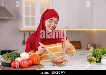 Donna musulmana che prepara una deliziosa insalata con verdure al tavolo bianco in cucina Foto Stock