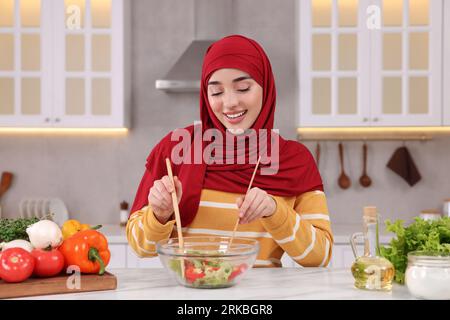 Donna musulmana che prepara una deliziosa insalata con verdure al tavolo bianco in cucina Foto Stock