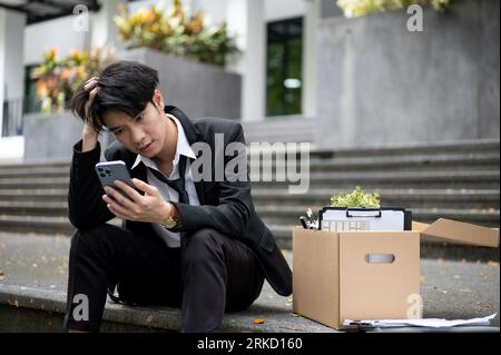 Stressato e turbato il giovane uomo d'affari asiatico o un lavoratore d'ufficio maschio che digita sul telefono mentre si siede su una scala con una scatola delle sue cose personali a poppa Foto Stock