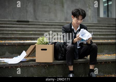 A frustrated and angry young Asian unemployed businessman is crumpling papers while sitting on the stairs in front of the corporate office with a box Foto Stock