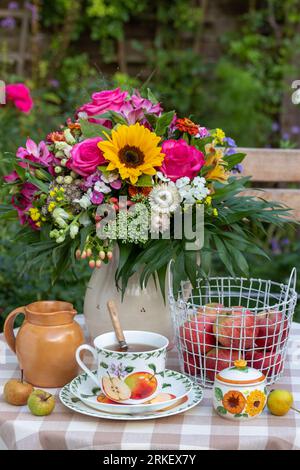 disposizione dei tavoli con tazza di tè, mele nel cestino e bouquet di fiori estivi Foto Stock