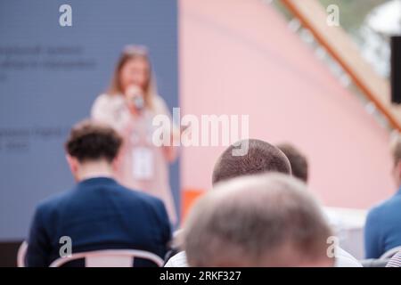 Vista posteriore di una folla anonima di persone che si riuniscono e ascoltano una speaker femminile con il microfono in mano Foto Stock