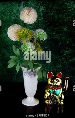 Fiori sintetici in un vaso decorato con smalto con neko maneki dorato che richiama il fortunato gatto su un tavolo in legno Foto Stock