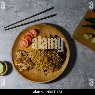 Mie goreng ayam geprek. Spaghetti fritti serviti su un piatto di legno accompagnato da pollo, pomodori freschi e cosparso di cipolle fritte Foto Stock