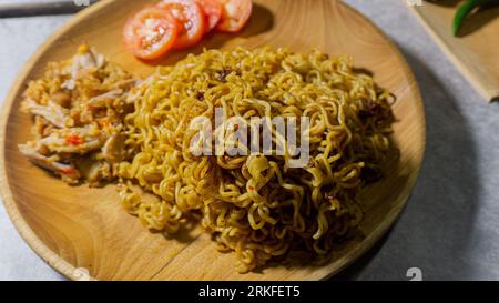 Mie goreng ayam geprek. Spaghetti fritti serviti su un piatto di legno accompagnato da pollo, pomodori freschi e cosparso di cipolle fritte Foto Stock