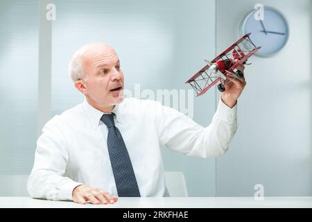 Older man, feeling youthful, plays with a red tin toy airplane on his work desk. Perhaps a visionary leader, he seeks inspiration through such leisure Stock Photo