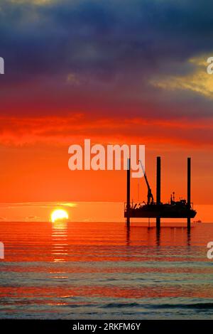 Carro di prova di perforazione con nuvole d'orizzonte dorate e cielo arancione e sole all'orizzonte al largo del Mare del Nord all'alba con cielo e mare dall'aspetto caldo Foto Stock