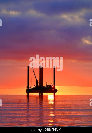 Carro di prova di perforazione con nuvole d'orizzonte dorate e cielo arancione e sole all'orizzonte al largo del Mare del Nord all'alba con cielo e mare dall'aspetto caldo Foto Stock