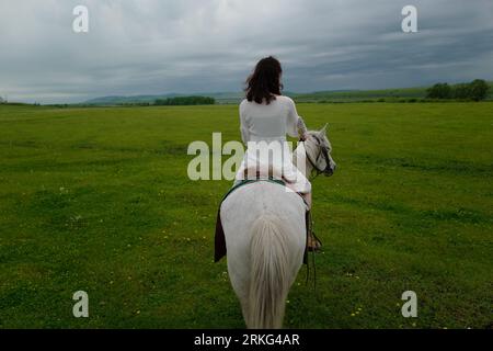 Una giovane donna sta cavalcando un cavallo in un campo aperto, guardando il cielo azzurro Foto Stock