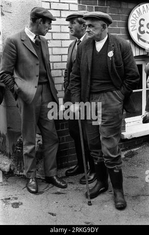 Agricoltori che discutono di un po' di affari nella città di mercato di Hatherleigh Devon 1970s nel Regno Unito. La vendita annuale di cavalli è appena finita, due uomini indossano abiti in tweet, un colletto e cravatta, saltatori (novembre) e scarpe in pelle lucida. Il terzo uomo con bastone da passeggio indossa una vecchia camicia senza colletto, un maglione e una vecchia giacca. Pantaloni a cordoncino e stivali wellington. Tutti e tre gli uomini indossano berretti piatti in tweed. Inghilterra 1973. HOMER SYKES Foto Stock