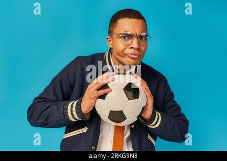 studente afro-americano serio in occhiali che tengono palla da calcio e guance gonfiabili sul blu Foto Stock