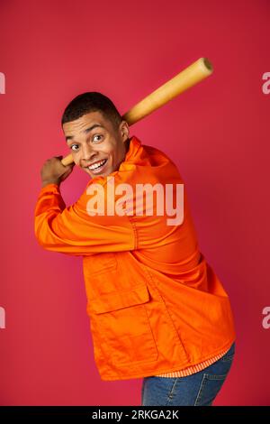 afroamericano energizzato con una camicia arancione che gioca a baseball su sfondo rosso Foto Stock