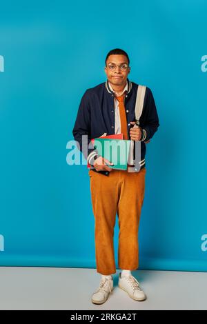 studente afroamericano annoiato con notebook e guance imbottite a zaino su sfondo blu Foto Stock