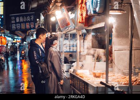 A young couple wearing raincoats and holding umbrellas while browsing the offerings of a street food stand on a wet and rainy evening Stock Photo